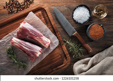 Raw Lamb Drumstick On White Cooking Paper And Wooden Cutting Board. Decorated With Herbs, Spices, Chef's Knife And Napkin. Overhead View.