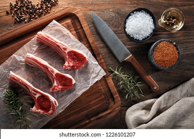Raw lamb rib chops meat on white cooking paper and wooden cutting board. Decorated with herbs, spices and chef's knife. Overhead view.