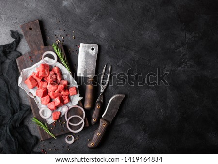 Raw lean diced casserole beef pork steak with vintage meat hatchet and fork with knife on stone background. Rosemary with red onion.