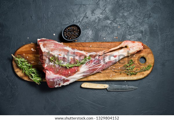 Raw leg of lamb on a\
wooden chopping Board. Rosemary, thyme, black pepper. Black\
background, top view