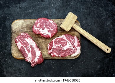 Raw Loin Beef Steaks, Wooden Hammer, Salt And Pepper Mill On Cutting Board. Beef Steaks BBQ Or Grilling, Overhead View. Raw Striploin Marbled Beef Steaks On Black Background, Top View.