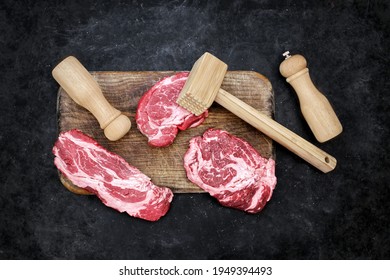 Raw Loin Beef Steaks, Wooden Hammer, Salt And Pepper Mill On Cutting Board. Beef Steaks BBQ Or Grilling, Overhead View. Raw Striploin Marbled Beef Steaks On Black Background, Top View.