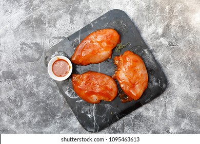 Raw Marinated Chicken Breast Fillets On Black Stone Plate Tray With BBQ Sauce In White Dish On Grey Concrete Background, Prepatation For Grilling Or Smoking, Top View, Overhead