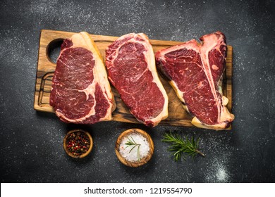 Raw meat beef steak. Black angus prime meat set - ribeye, striploin, t-bone steaks on cutting board. Top view on black table. - Powered by Shutterstock