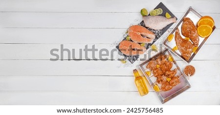 Raw meat and fish arranged on a white table, featuring a red fish steak, sea bass carcass, marinated chicken breast, and duck shank. Top view