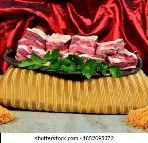 Raw meat ribs on a black clay plate, gold and red silk fabric background. Raw Organic Beef Short Ribs decorated with green persil leaves