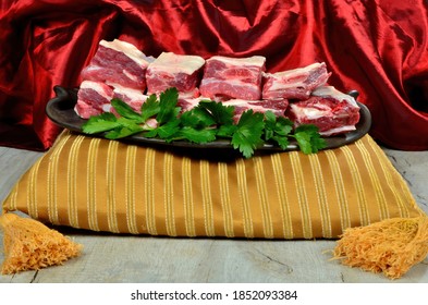Raw meat ribs on a black clay plate, gold and red silk fabric background. Raw Organic Beef Short Ribs decorated with green persil leaves