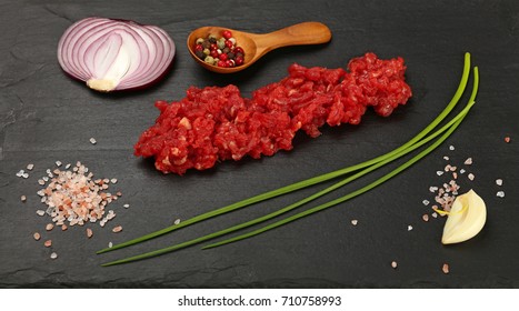 Raw minced beef red meat cutlet, spices, peppercorn in wooden scoop, spring green chive, onion, garlic cloves and Himalayan salt on black slate board, close up, high angle view - Powered by Shutterstock