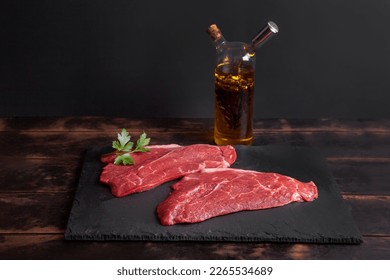 Raw minute steak of marbled beef with a parsley leaf on a stone slate board, glass bottle with vegetable oil and balsamic vinegar.