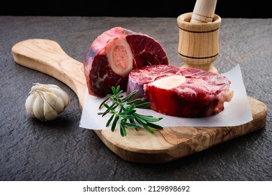 Raw osso buco steak lies on waxpaper. Fresh veal shank meat cut with marrow bone served at wooden board decorated with garlic bulb and rosemary