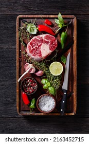 raw osso buco, veal shank steak on wood rustic cutting board with peppercorns, fresh thyme, chili  peppers, garlic and lime on dark wood table, vertical view