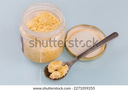 Raw partially crumbled solid cocoa butter in the small glass jar with removed lid and pieces in a tea spoon on a blue surface