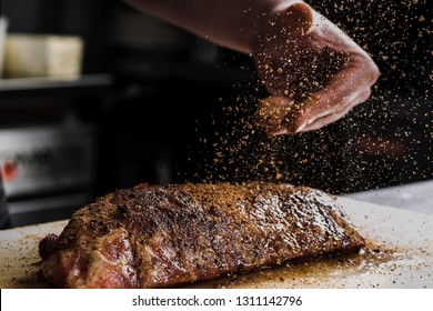 Raw piece of meat, beef ribs. The hand of a male chef puts salt and spices on a dark background, close-up. - Shutterstock ID 1311142796