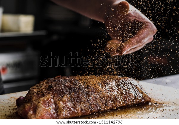 Raw piece of meat,\
beef ribs. The hand of a male chef puts salt and spices on a dark\
background, close-up.