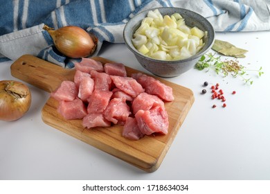 Raw Pieces Of Pork Meat On A Kitchen Board And Ingredients Like Onions, Herbs And Spices On A White Table To Cook Stew, Goulash Or Ragout, Selected Focus, Narrow Depth Of Field