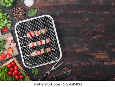 Raw pork kebab with paprika on disposable coal bbq grill with fresh vegetables on wooden background with fork and knife. Salt and pepper with lettuce and paprika.