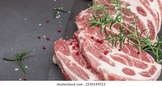 Raw pork neck meat. Chop steak, red peppercorn, garlic cloves, sea salt and fresh rosemary. Trendy stand, black background, flat lay, banner format - Powered by Shutterstock