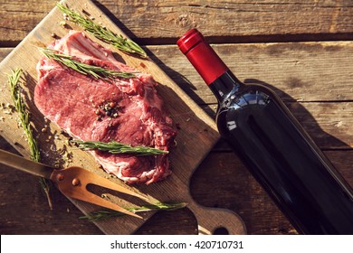 Raw Pork Steak With Bottle Of Red Wine, Meat Fork And Rosemary On Wooden Background