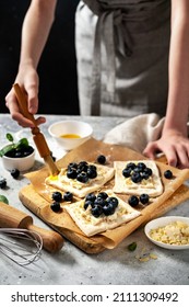 Raw puff pastry with cheese cream and blueberries. The process of making delicious sweet buns or mini pies. Greasing the edges of the baking dough with egg. Hands in the frame	