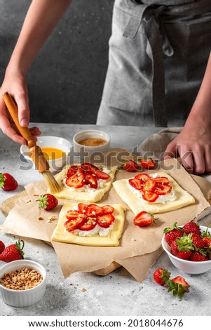 Raw puff pastry with cheese cream and strawberries. The process of making delicious sweet buns. Greasing the edges of the baking dough with egg. Hands in the frame