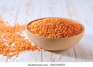 Raw red lentils are scattered out of the wooden bowl on a light table, selective focus.