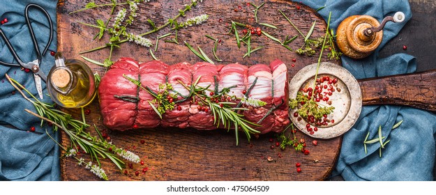 Raw roast beef  with herbs tied with a rope with cooking ingredients, oil  and spices on rustic background, top view, banner
