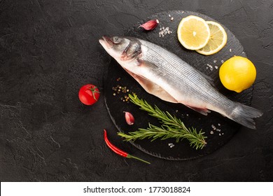 Raw sea bass with fresh rosemary,lemon,cherry pepper,garlic and tomato on stone plate on black concrete background.Top vie,copy space.