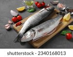Raw seabass with ingredients and seasonings like basil, lemon, salt, pepper, cherry tomatoes and garlic on wooden board on gray background