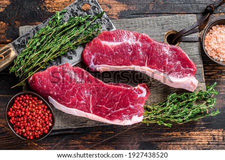 Raw sirloin beef meat steak on a wooden cutting board with herbs. Dark wooden background. Top view.