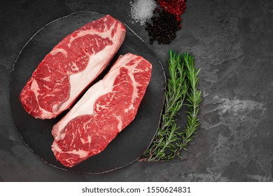 Raw Striploin Beef Steak With Rosemary, Salt And Pepper On Round Slate On Stone Table. Top View