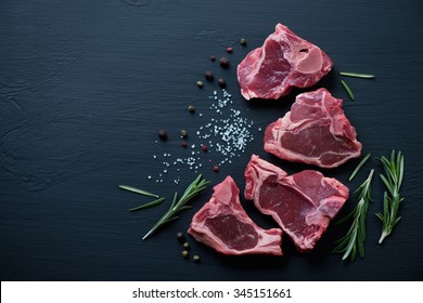 Raw T-bone lamb steaks with seasonings on a black wooden surface