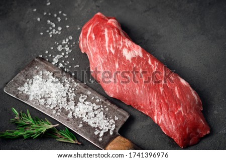 Raw tenderloin of beef or skirt steak with a hatchet for meat, salt and rosemary on a dark stone background, close up