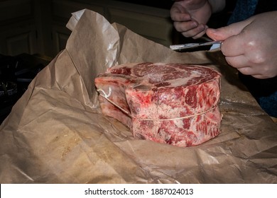Raw two bone stnading rib beef roast on butchers paper with pepper on it being sprinked with salt from an silver oar spoon by hands
