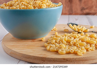 Raw or uncooked Elbow Pasta in a blue bowl and on a chooping wooden board witrh a spoon on the side and a wooden wall in the background