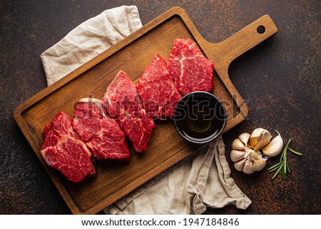 Raw uncooked meat beef cubes for stew on wooden cutting board with seasonings, olive oil, garlic and fresh rosemary on brown rustic stone background 