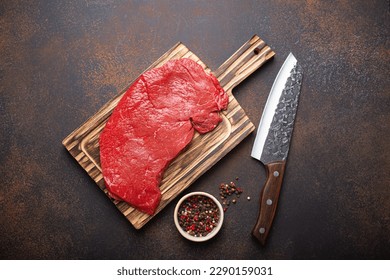 Raw uncooked top round beef steak on wooden cutting board with big kitchen knife and pepper on dark brown rustic stone background top view, cooking meat steak