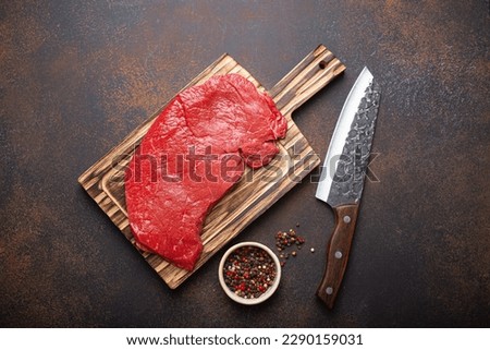 Raw uncooked top round beef steak on wooden cutting board with big kitchen knife and pepper on dark brown rustic stone background top view, cooking meat steak