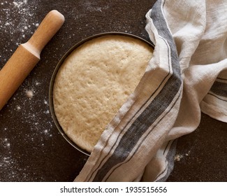 Raw yeast dough resting and rising in large metal bowl covering with linen towel on dark background. Wooden rolling pin and flour scattering on table