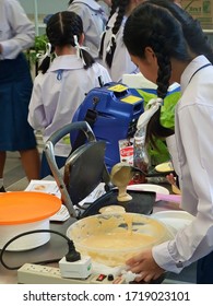 Rayong, Thailand - 6th September, 2018: A Thai High-school Girl In Uniform Pours Mixture Onto The Hotplate Of A Machine To Cook Pancakes During A Cookery Lesson, Eastern Thailand.