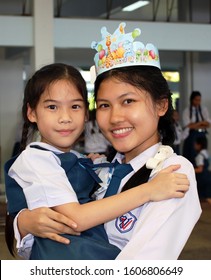 Rayong, Thailand - 7th September, 2017: An Older Female Teenage High-school Student Lifts Up And Holds A Younger Primary Girl At A Party At A School, Eastern Thailand.