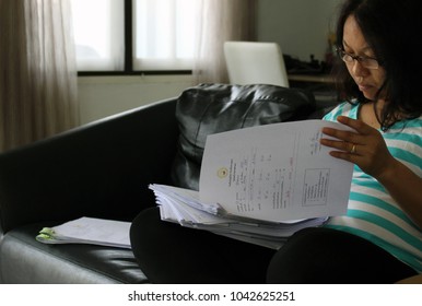 Rayong, Thailand - March 10th, 2018: A Teacher Working From Home Sits On A Sofa Grading A Pile Of School Student Exam Papers