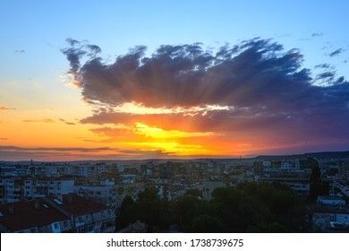 The rays of the setting sun break through a large cloud while sun goes down over the horizon. Golden blue evening sky over a small town. Summer skyscape.