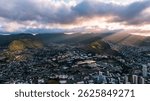 Rays of setting sun go through the grey clouds lighting the mountains. Drone flight above the dense urban landscape of Honolulu, Hawaii, USA.