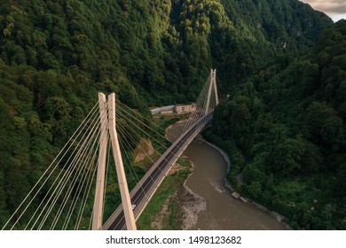 The rays of the setting sun illuminate the cable-stayed automobile bridge surrounded by mountains covered with forests/ Aerial view from drone