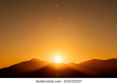 rays of the sun setting behind a mountain with a completely clear, orange sky at sunset