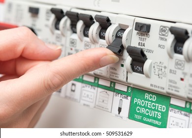 An RCD circuit breaker board displays many switches. Most are in the ON position, but one is switched down to OFF. It is the circuit for the lighting. A finger is about to turn it back on