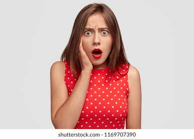 Reaction and emotions concept. Shocked emotional gorgeous young female person exclaims with stupefaction, hears bad negative news, wears red polka dot dress, stands against white background.