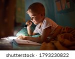 Reading book and using flashlight. Young boy in casual clothes lying down near tent at evening time.