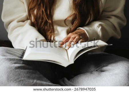 Reading a book while sitting on the bed. Cozy lifestyle with woman in casual clothes. Holding open book on knees. Relaxing leisure time indoors. Simple everyday moment of calm.