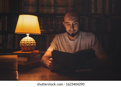 Reading And Education. A Man In The Library Reading A Book In The Evening By The Light Of A Lamp. In The Background, A Rack Of Books. Dark Room